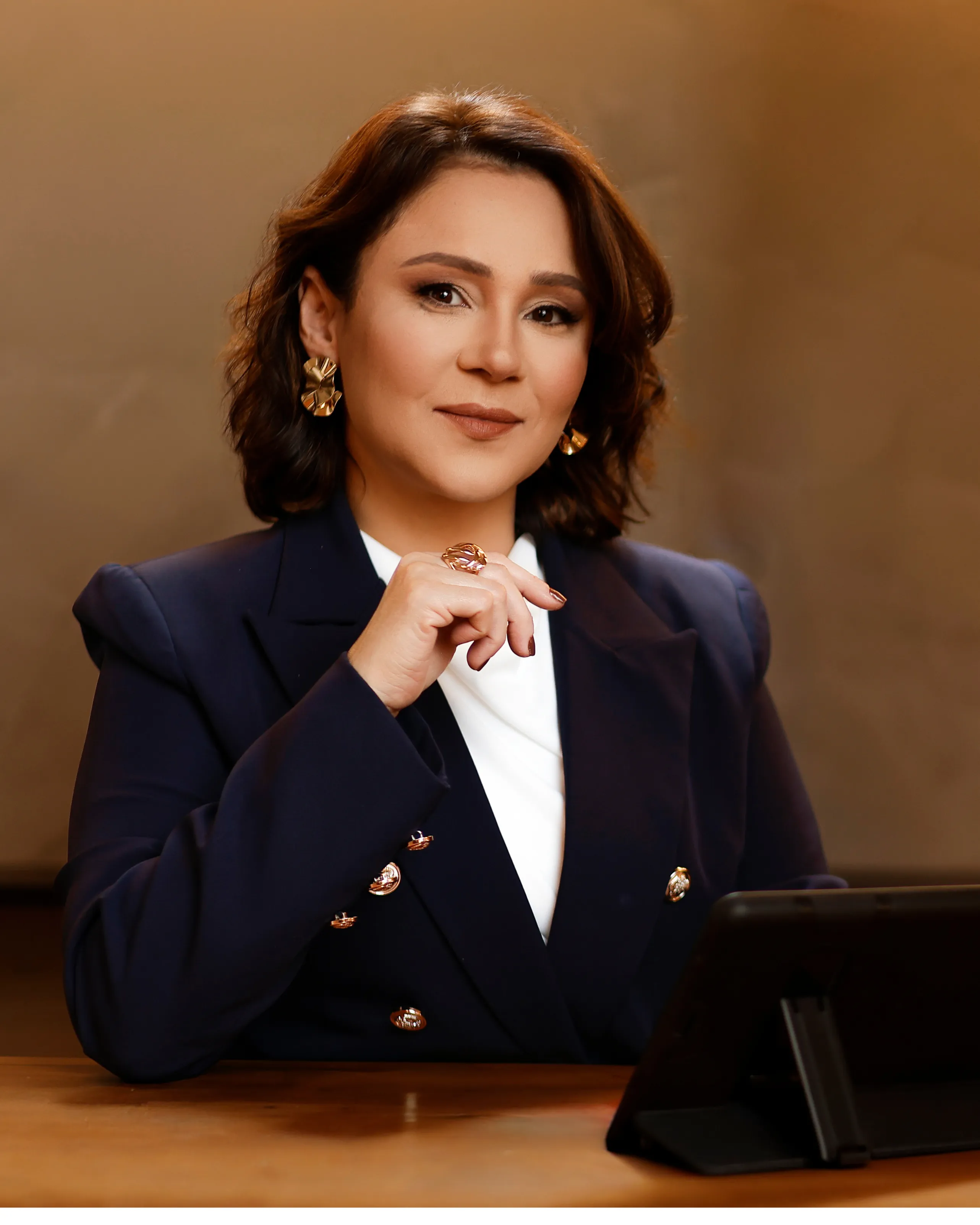 Katiuscia Silva, de blazer azul-marinho, sentada à mesa com a mão no queixo, ao lado de um tablet, em postura confiante.