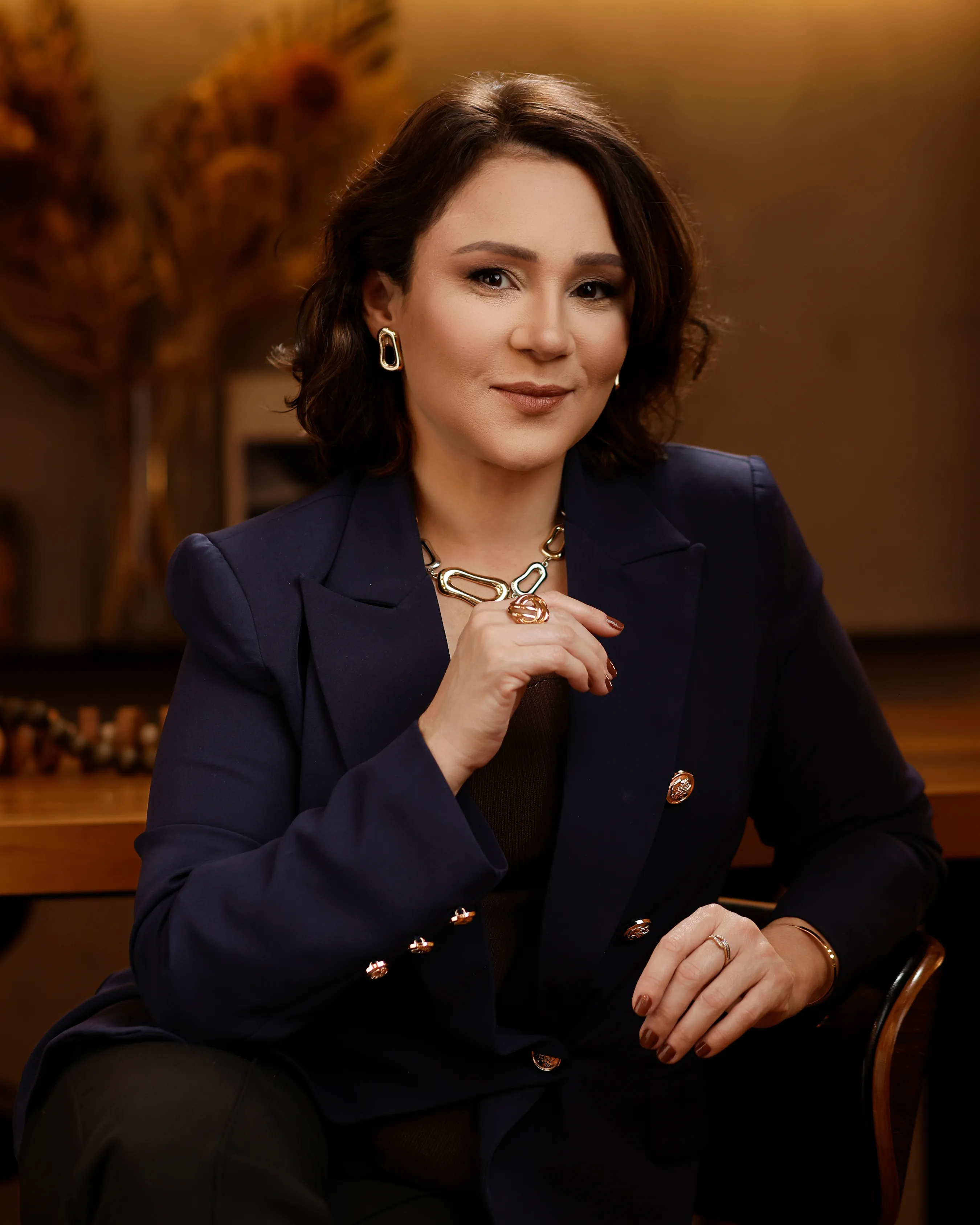 Retrato de Katiuscia Silva, de blazer azul-marinho, sentada com a mão próxima ao colar e expressão serena e confiante.
